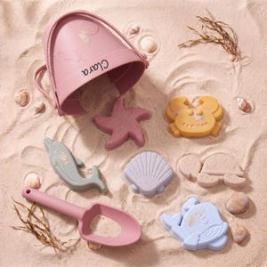 Pode incluir: Um balde de areia rosa com o nome "Clara" impresso, rodeado por moldes de areia de uma estrela do mar, caranguejo, golfinho, concha, tartaruga, baleia e uma pá rosa. Os brinquedos de areia são em várias cores pastel.