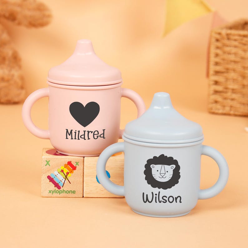 Puede incluir: Dos vasos con boquilla personalizados para beb&eacute;s. Uno es rosa claro con un coraz&oacute;n negro y el nombre "Mildred". El otro es azul claro con un dise&ntilde;o de le&oacute;n y el nombre "Wilson". Ambos tienen asas.