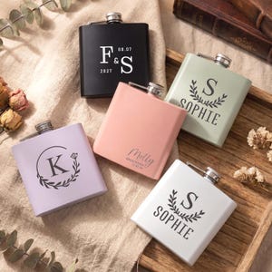 May include: A selection of personalised hip flasks in shades of black, pink, lavender, green, and white. Each flask displays custom text, including initials, names, and dates, with floral designs. The flasks have a silver cap.