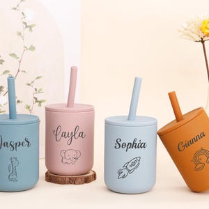 May include: Four colorful silicone cups with lids and straws. Each cup is a different color: blue, pink, light blue, and orange. The cups are personalized with names and small illustrations: Jasper, Cayla, Sophia, and Gianna. The cups are designed for children.
