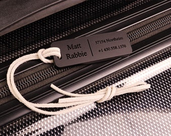 May include: Black luggage tag with the name "Matt Rabbie", address "37154 Northeim", and phone number "+1 430.558.1376" engraved on it. The tag is attached to a black suitcase with a white leather strap.