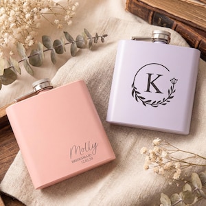 May include: Two personalised hip flasks. One is pale pink with the name "Molly" and the text "Bridesmaid 12.02.30". The other is lavender with a black floral wreath and the letter "K". Both have a silver cap.