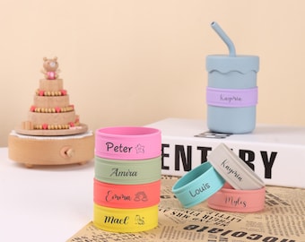 May include: A set of colorful silicone wristbands with personalized names, including Peter, Amira, Emora, Macl, Louis, Kaypria, and Myles. The wristbands are stacked on top of each other, with the names facing outward. There is also a light blue silicone cup with a straw and a purple silicone band with the name Kaypria on it.