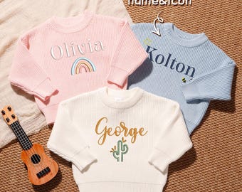 May include: Three personalized children's sweaters in pastel colors: pink, light blue, and white. Each sweater features a custom name and icon. The pink sweater has the name "Olivia" with a rainbow, the blue sweater has "Kolton", and the white sweater has "George" with a cactus.