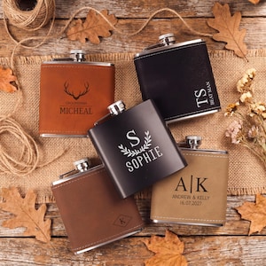 May include: An assortment of personalized flasks in various colors, including brown, black, and tan. Each flask features custom engravings with names, initials, and dates. The flasks are arranged on a rustic wooden surface with autumn leaves.