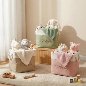Peut inclure: Trois bacs de rangement en tissu beige, vert et rose, chacun avec un dessus blanc et des poignées en corde. Les bacs sont remplis d'animaux en peluche, de couvertures et de jouets. Le bac vert porte le nom "Apollo" brodé. Le bac rose porte le nom "Veirity" brodé.