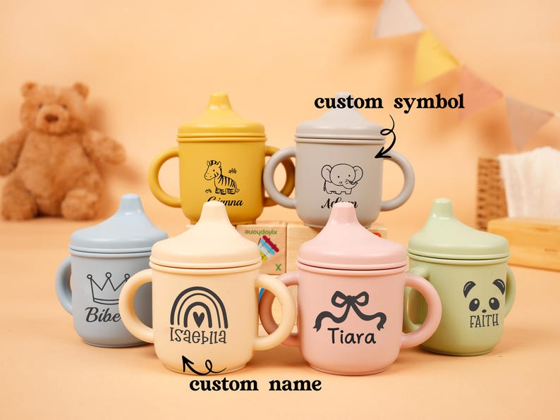 Puede incluir: Una colecci&oacute;n de coloridas tazas para beb&eacute;s con asas y tapas. Cada taza presenta un dise&ntilde;o diferente, incluyendo una cebra, un elefante, un panda, una corona, un arco&iacute;ris y un lazo. Las tazas son en tonos amarillo, gris, rosa, verde y azul.