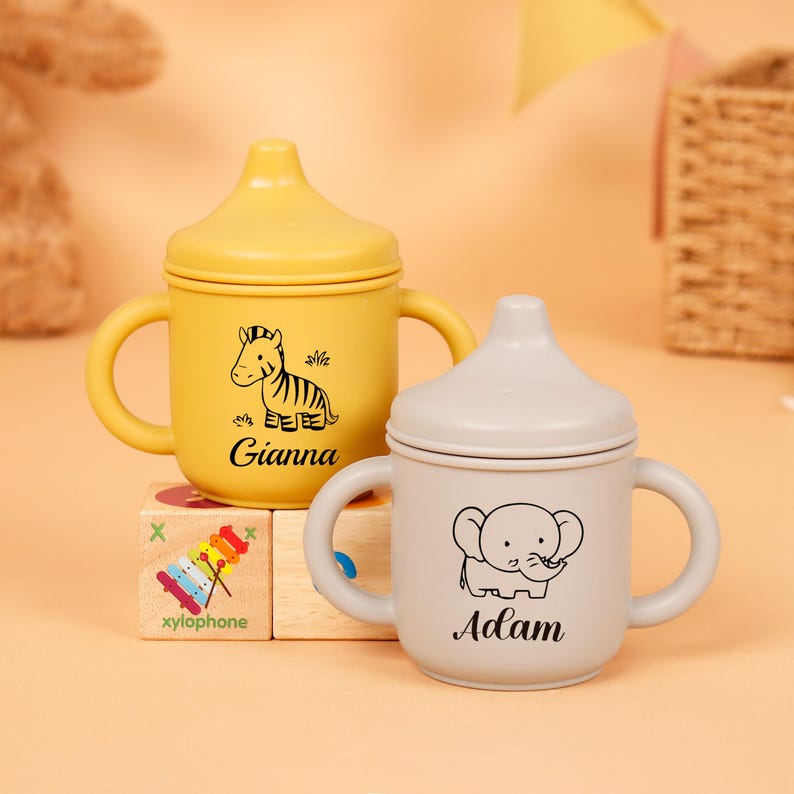 Puede incluir: Dos vasos para beb&eacute;s con tapa. Uno es amarillo con un dibujo de cebra y el nombre "Gianna". El otro es gris con un dibujo de elefante y el nombre "Adam". Ambas tazas tienen asas.