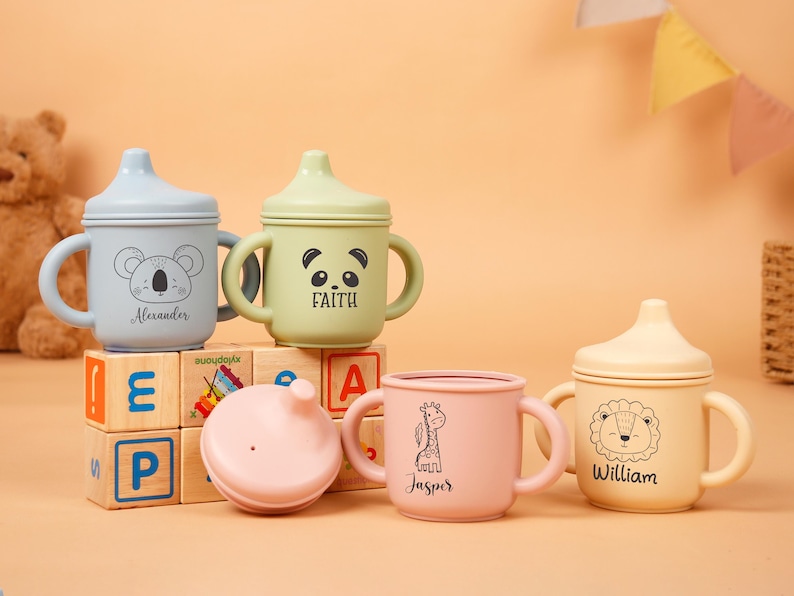 Puede incluir: Una colecci&oacute;n de vasos con boquilla para beb&eacute;s en colores pastel, cada uno con una ilustraci&oacute;n de animal &uacute;nica y el nombre de un ni&ntilde;o. Las tazas tienen asas y tapas, y est&aacute;n dispuestas sobre bloques de madera. Un oso de peluche y banderas decorativas est&aacute;n en el fondo.