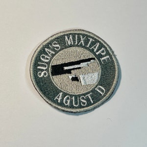 May include: A green embroidered patch with the text "Sugas Mixtape August D" in white lettering. The patch features a black and white graphic of a microphone.