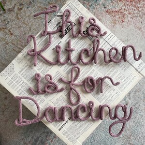May include: A close-up of a pink knitted phrase that reads "This kitchen is for dancing" on a newspaper background.