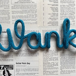 May include: The word "Thank" spelled out in blue yarn, with a knitted texture. The yarn is arranged in a rope-like shape, creating a three-dimensional effect. The background is a page from a knitting pattern book.