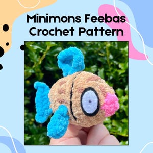 Puede incluir: Un juguete de pez Feebas tejido a ganchillo con aletas azules y una aleta cefálica azul. El pez tiene un cuerpo marrón con manchas marrones, una boca rosa y un ojo blanco y negro. El texto "Minimons Feebas Crochet Pattern" está en la parte superior.
