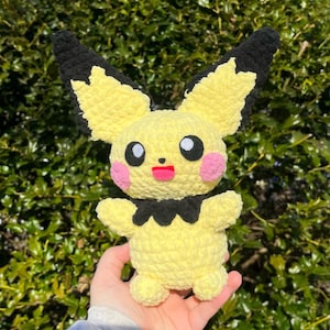 May include: A handmade yellow crochet Pichu plushie with black-tipped ears, black eyes, pink cheeks, and a red mouth. The plushie has small arms and a black collar. The background is a green bush.
