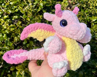 Puede incluir: Un dragón rosa de ganchillo hecho a mano con alas amarillas y rosas, detalles blancos y ojos azules. El peluche tiene una cola larga y pequeños cuernos. El dragón es un juguete suave y caprichoso.