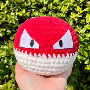 Può includere: Una palla all'uncinetto rossa e bianca con occhi triangolari bianchi e pupille nere. La metà inferiore è bianca e la parte superiore è rossa. Gli occhi sono in feltro bianco. La palla è rotonda e morbida.