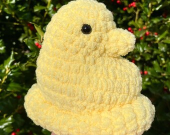 Puede incluir: Un pollito de ganchillo amarillo hecho a mano con un ojo negro. El pollito es suave y esponjoso, con un cuerpo redondeado y un pico pequeño. El pollito se sostiene en una mano sobre un fondo de follaje verde.