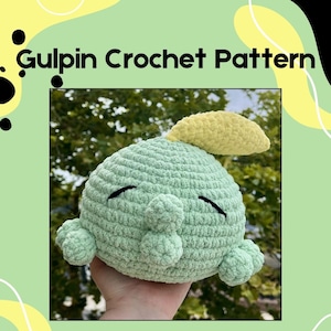 Puede incluir: Un juguete de peluche tejido a ganchillo en tonos verdes y amarillos, que se asemeja a una criatura caprichosa. El juguete tiene un cuerpo redondeado, pequeños apéndices y ojos negros cerrados. El texto "Gulpin Crochet Pattern" se muestra en la parte superior.