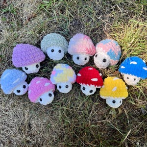 May include: A collection of handmade crochet mushroom figures in various colors, including purple, pink, blue, red, and yellow. Each mushroom has a white stem and black button eyes, arranged on a bed of grass.