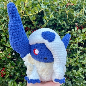 May include: A handmade crochet plush toy in the shape of a character with blue and white details. The toy has large blue ears, a white head with blue accents, and a red eye. The plush is made with soft yarn and has a cute, friendly appearance.