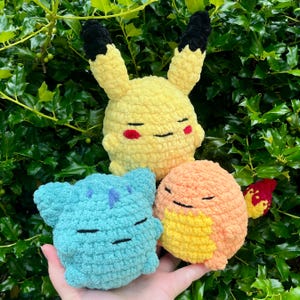 Puede incluir: Peluche de ganchillo de Pikachu, Bulbasaur y Charmander. Pikachu es amarillo con orejas con puntas negras y mejillas rojas. Bulbasaur es azul y Charmander es naranja. Los tres tienen ojos y bocas cosidas en negro.