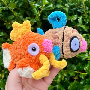 Puede incluir: Dos juguetes de peces de ganchillo. Uno es naranja con detalles amarillos, blancos y rosas. El otro es marrón con detalles azules y rosas. Ambos tienen ojos grandes y redondos. Los juguetes se sostienen en una mano sobre un fondo de hojas verdes.