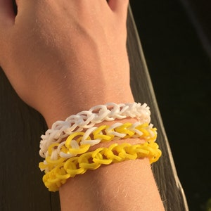 May include: A stack of three bracelets on a person's wrist. The bracelets are made of yellow and white rubber bands. The bracelets are in a chain link pattern.