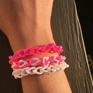 May include: A stack of three bracelets on a person's wrist. The top bracelet is pink, the middle bracelet is pink and white, and the bottom bracelet is white.