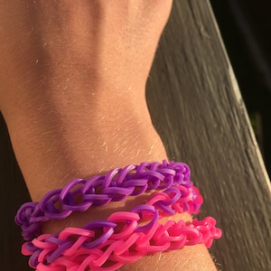 May include: Two colorful rubber band bracelets on a person's wrist. The top bracelet is purple and the bottom bracelet is pink.