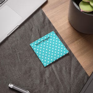 Op de afbeelding: Een vierkante, turquoise plaknotitie met witte stippen. De notitie heeft de tekst "Don't forget..." in het zwart geschreven. De notitie ligt op een donkergrijze ondergrond, in de buurt van een laptop en een pen.