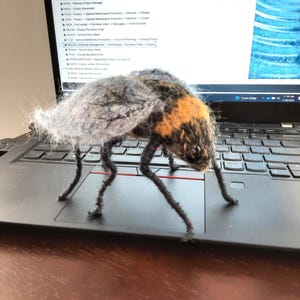 May include: A handcrafted fly model with a grey body, an orange and black head, and long, dark legs. The fly is positioned on a laptop keyboard, with a computer screen showing text and a blue abstract design in the background.