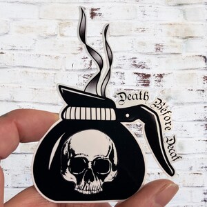 May include: A black and white sticker depicting a coffee pot with a skull and the words "Death Before Decaf". The sticker has a steam design rising from the top. The sticker is held by a hand.