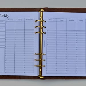 May include: Open brown leather planner with gold-colored rings and a weekly schedule insert. The insert is white with black text, including the word "Weekly" at the top. The planner has a zipper pocket and a snap closure.