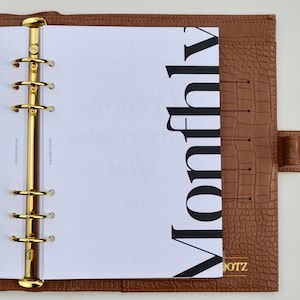 May include: A brown leather planner with gold hardware and the word "Monthly" printed in large black letters on a white page. The planner has a textured, crocodile-like pattern and the brand name "DOTZ" in gold.