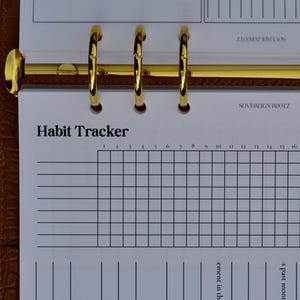 May include: A gold-colored binder ring mechanism holds a white habit tracker sheet. The sheet is labeled "Habit Tracker" and includes a grid for tracking habits. The binder is set in a brown leather-like cover.