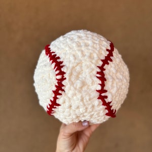 May include: A handmade, cream-coloured baseball with red stitching. The ball is made of a soft, textured material, and the red stitching is clearly visible. This baseball is a decorative item.