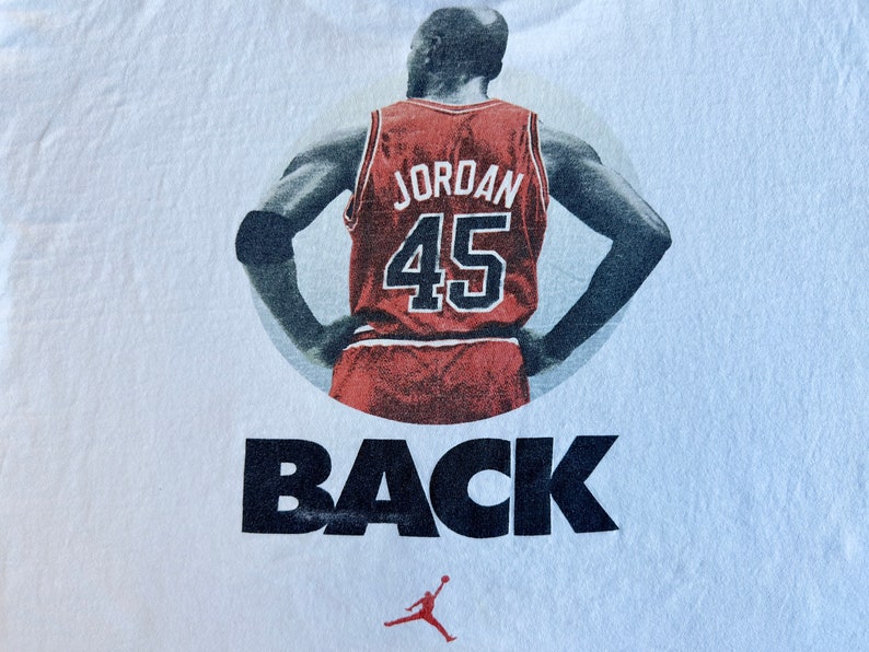Pode incluir: Camiseta branca com uma imagem em preto e branco de um jogador de basquete vestindo uma camisa vermelha com o n&uacute;mero 45 e a palavra "JORDAN" nas costas. O texto "BACK" &eacute; impresso abaixo da imagem. Uma pequena silhueta de jogador de basquete vermelho e preto &eacute; impressa abaixo do texto.