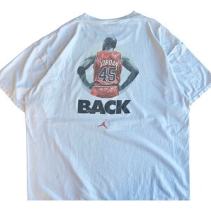 Pode incluir: Camiseta branca com um gr&aacute;fico de Michael Jordan vestindo uma camisa dos Chicago Bulls vermelha e preta com o n&uacute;mero 45 nas costas. O texto "BACK" est&aacute; impresso abaixo do gr&aacute;fico.