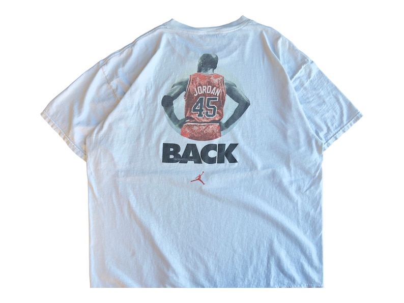 Pode incluir: Camiseta branca com um gr&aacute;fico de Michael Jordan vestindo uma camisa dos Chicago Bulls vermelha e preta com o n&uacute;mero 45 nas costas. O texto "BACK" est&aacute; impresso abaixo do gr&aacute;fico.