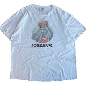 Pode incluir: Uma camiseta branca vintage com uma imagem desbotada de Michael Jordan jogando basquete. A camiseta tem o texto "JORDAN'S" impresso abaixo da imagem.