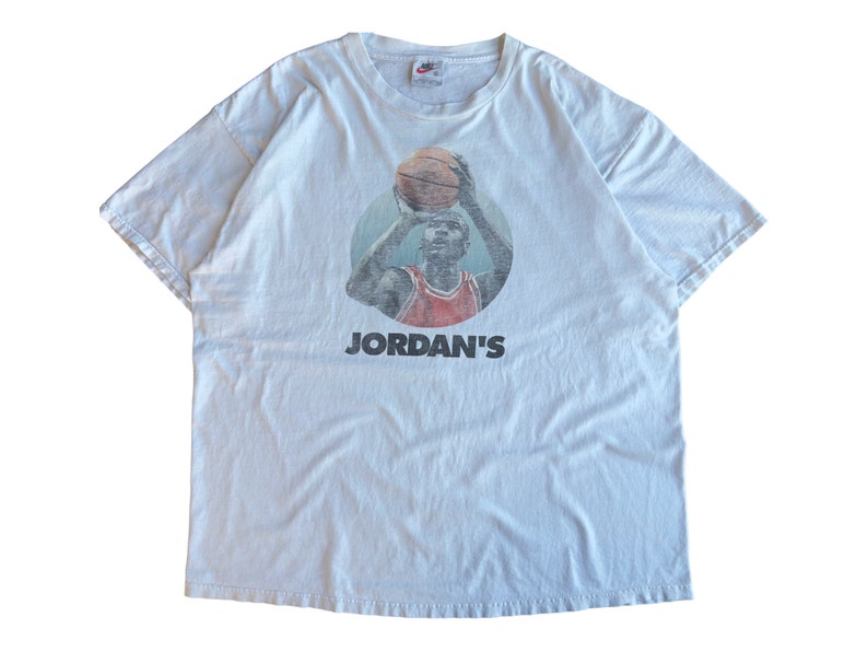Pode incluir: Uma camiseta branca vintage com uma imagem desbotada de Michael Jordan jogando basquete. A camiseta tem o texto "JORDAN'S" impresso abaixo da imagem.