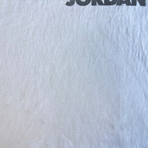 Pode incluir: Uma camiseta branca lisa com a palavra "JORDAN'S" impressa em preto no topo.