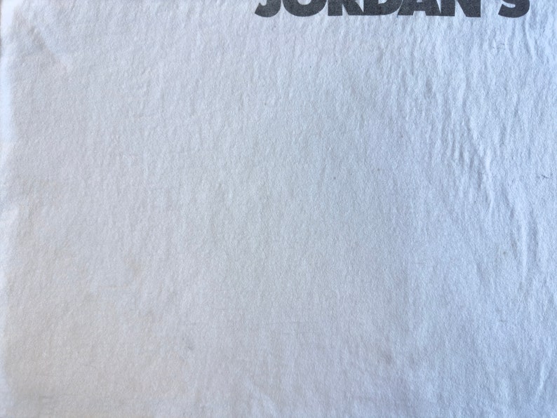 Pode incluir: Uma camiseta branca lisa com a palavra "JORDAN'S" impressa em preto no topo.