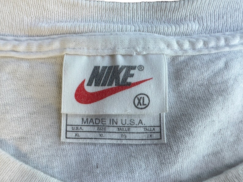 Pode incluir: Etiqueta de camiseta Nike branca com logo swoosh vermelho e o texto "MADE IN U.S.A.". A etiqueta tamb&eacute;m mostra o tamanho "XL" em tamanhos americano e internacional.