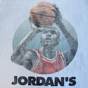 Pode incluir: Uma camiseta branca com uma imagem vintage de um jogador de basquete com um jersey vermelho e branco, segurando uma bola de basquete acima de sua cabe&ccedil;a. O texto "JORDAN'S" est&aacute; impresso em preto abaixo da imagem.