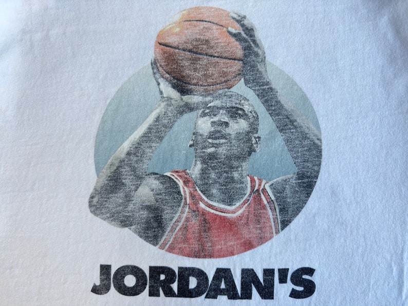 Pode incluir: Uma camiseta branca com uma imagem vintage de um jogador de basquete com um jersey vermelho e branco, segurando uma bola de basquete acima de sua cabe&ccedil;a. O texto "JORDAN'S" est&aacute; impresso em preto abaixo da imagem.