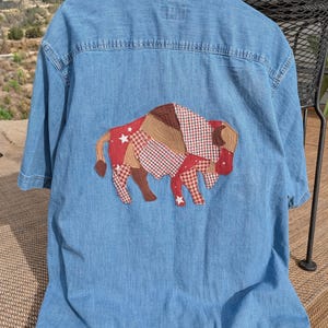 May include: Light blue denim shirt with short sleeves. The back of the shirt features a patchwork bison design in red, brown, and white fabrics. The bison has a star pattern and a gingham pattern.
