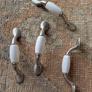 May include: Four cabinet pulls with a white ceramic center and brushed nickel finish. The pulls have a curved design and are arranged on a patterned rug. These decorative pulls are suitable for drawers and cabinets.