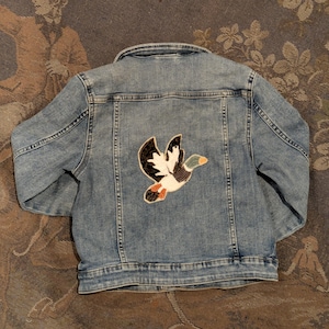 May include: A light blue denim jacket featuring a long-sleeved design with a fold-over collar and a button-up front. The back panel displays a large, detailed embroidered patch of a flying mallard duck with black, white, green, and orange colours.