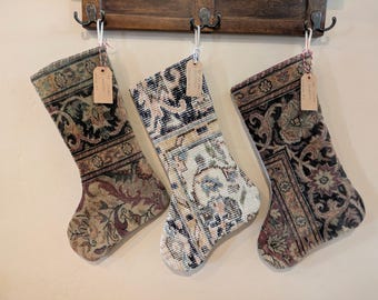 May include: Three Christmas stockings with intricate, patterned designs suspended from a wooden rack. The stockings display a mix of colours, including beige, black, and blue, with floral and geometric patterns. Each stocking has a small tag attached.